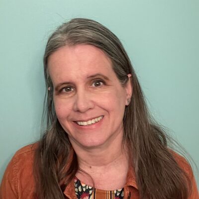 Headshot of Amy Barnes, a Mom&eacute; contributor, smiling in a rust corduroy jacket over a floral top against a mint green wall.
