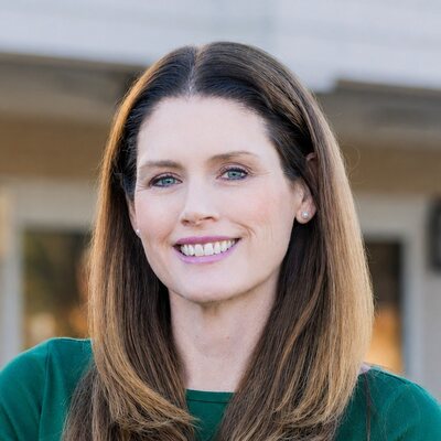 Headshot of Kelly O&rsquo;Lone, a Mom&eacute; contributor, smiling in a green boat-neck top with long brown hair, photographed outdoors.