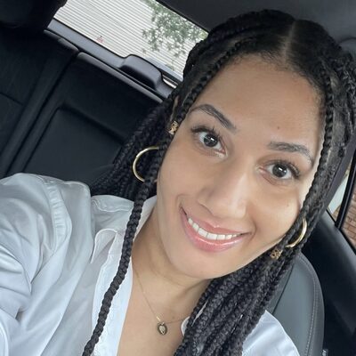Headshot of Princess Gabbara, a Mom&eacute; contributor, smiling in a white button-down shirt with long braided hair and gold hoop earrings.