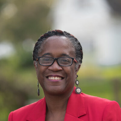 Headshot of Sheryl Nance-Nash, a Mom&eacute; contributor, smiling in a red blazer and black top with dangling earrings, photographed outdoors.