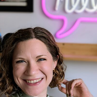 Headshot of Tassia O&rsquo;Callaghan, a Mom&eacute; contributor, smiling in a sage green top in front of a pink neon &lsquo;hello&rsquo; sign.