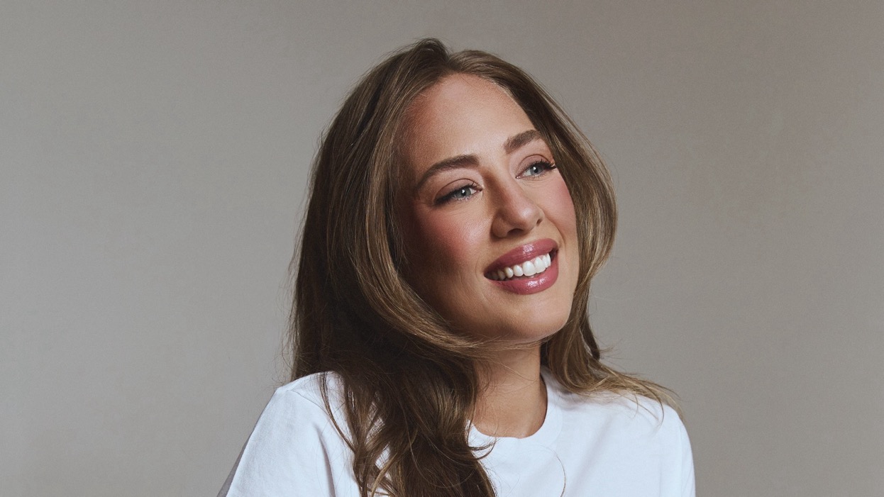 Amanda Hirsch, in a white crewneck T-shirt with long, wavy highlighted hair, smiles broadly and looks off to the side against a soft gray backdrop.