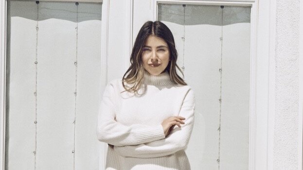Daniella Monet stands with her arms crossed in front of white French doors, wearing a cream turtleneck sweater.