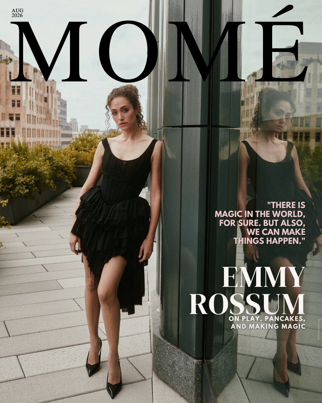 Momé August 2026 cover — Emmy Rossum in a black corseted tulle dress leaning against a dark glass column on a city rooftop, beneath the cover lines 'There is magic in the world, for sure. But also, we can make things happen.' and 'Emmy Rossum on play, pancakes, and making magic.'