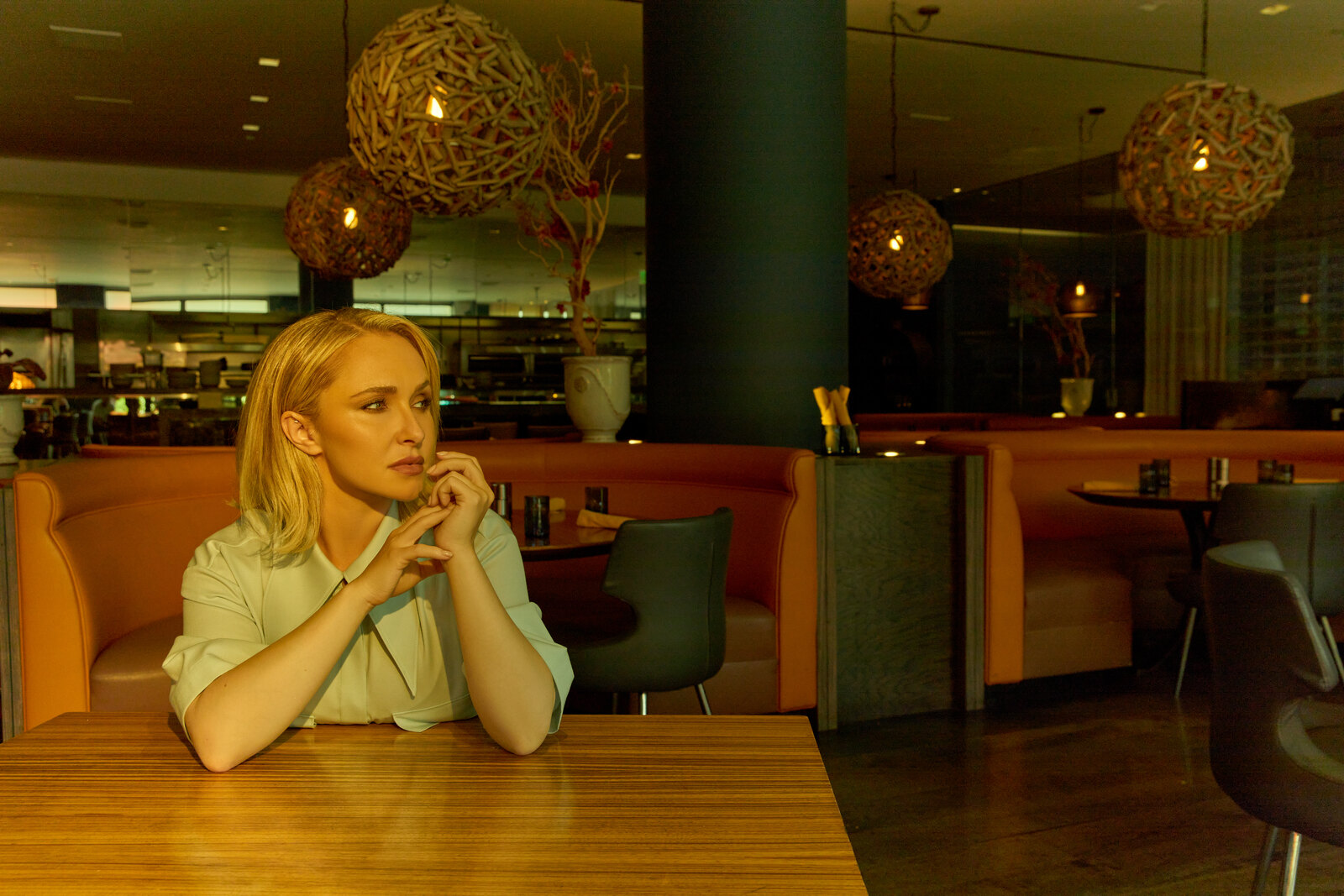 Hayden Panettiere seated at a restaurant table in a pale shirt, chin resting on her folded hands, looking thoughtfully into the distance under warm pendant lighting.