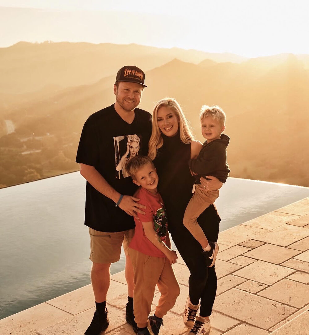 Heidi Montag, Spencer Pratt, and their two young sons stand together beside an infinity pool at golden hour, the hills glowing behind them.