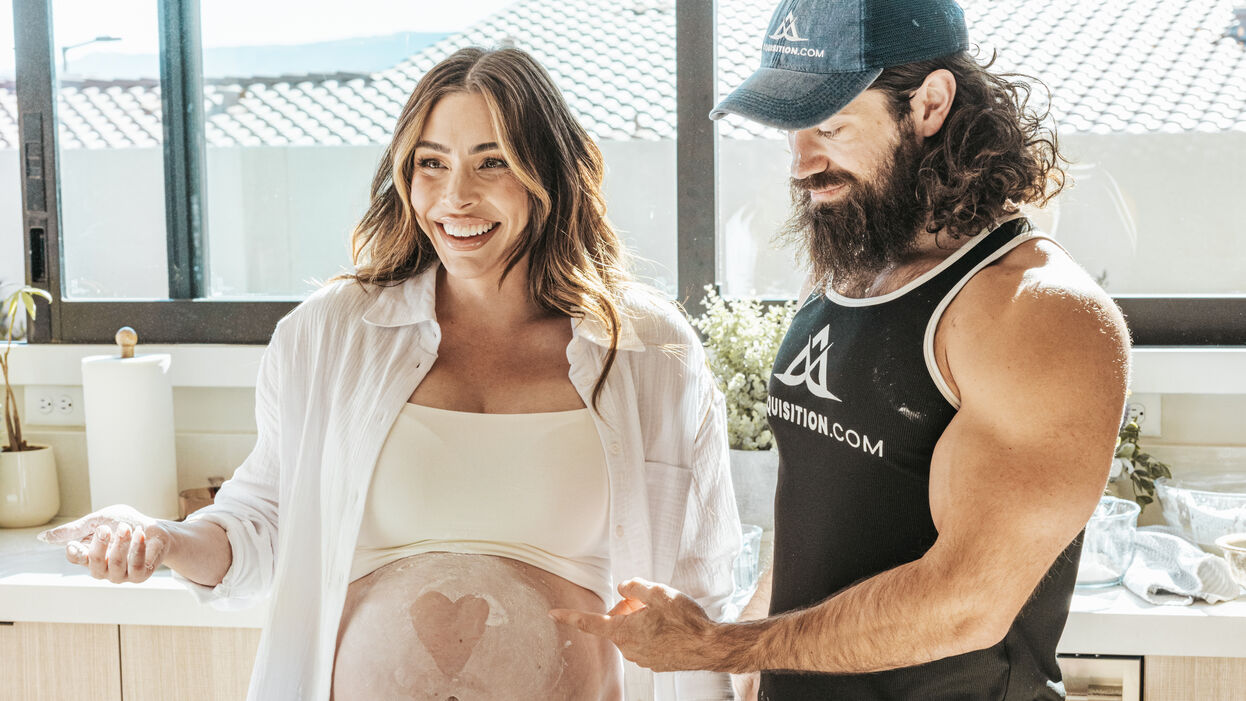 Leila Hormozi, in an open white shirt over a cream crop top with a heart painted on her bare pregnant belly, laughs beside her husband, Alex, in a cap and black tank top, as they bake together at a sunlit kitchen counter.