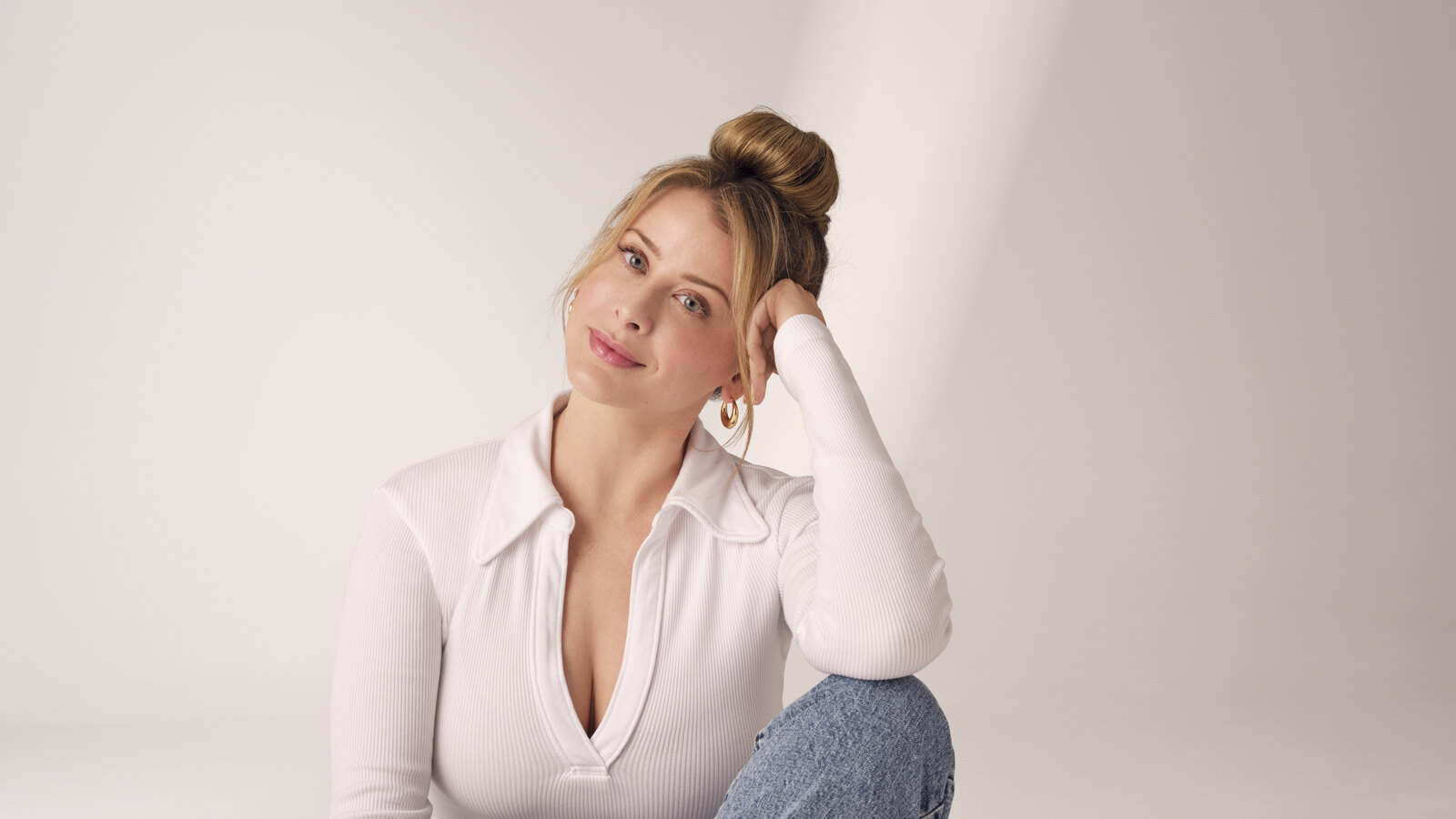 Lo Bosworth sits cross-legged on the floor of a pale studio in a white ribbed collared top and light-wash jeans, resting her head on one hand and looking directly at the camera.