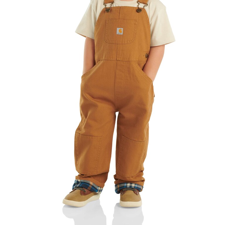 Carhartt Brown canvas bib overalls worn over a cream t-shirt by a small child, blue plaid flannel lining turned up at the cuffs above tan sneakers.