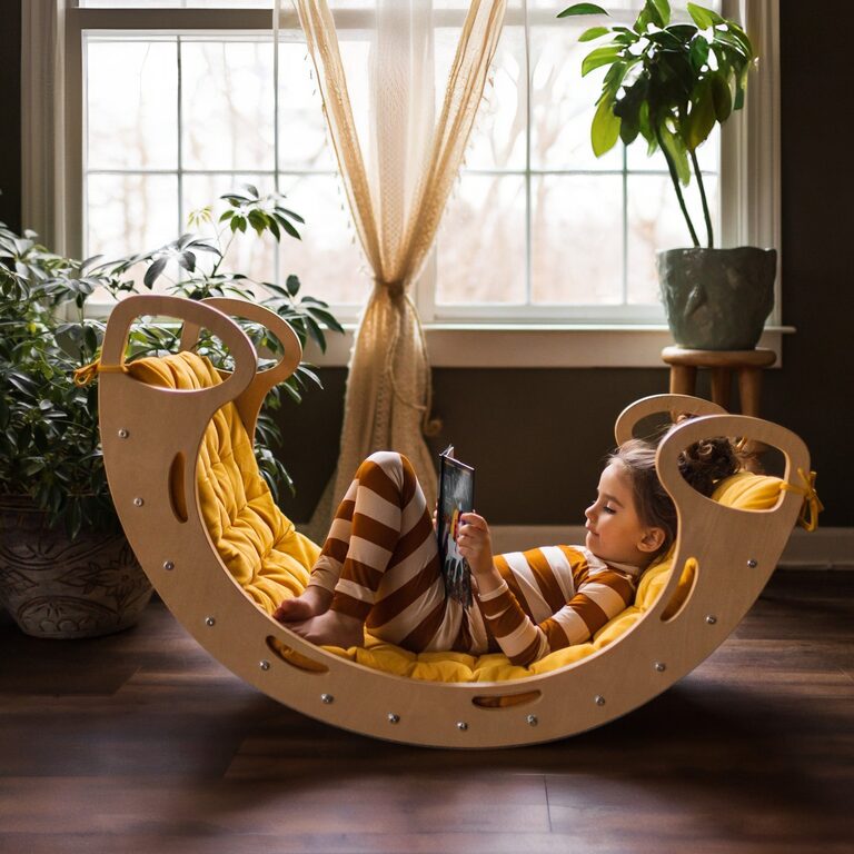 A child in striped pajamas lies reading a book in a Cassaro Jumbo Waldorf Rocker in natural birch, a curved wooden rocker with looped handles lined with a marigold-yellow cushion, in front of a bright window and houseplants.