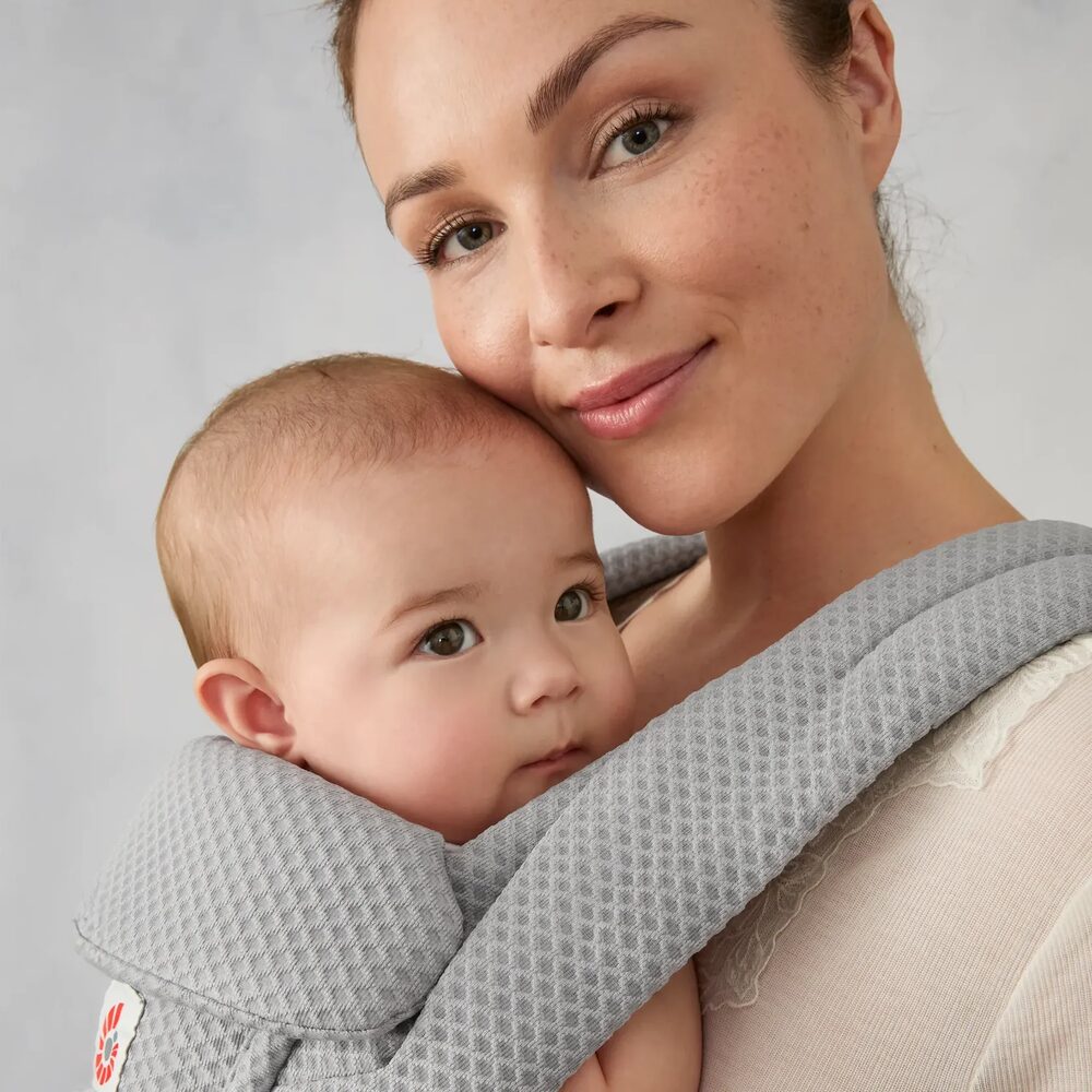 A smiling mother holds her baby close in a soft grey Ergobaby Omni Deluxe carrier.