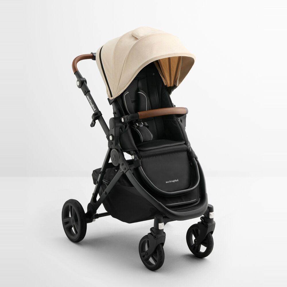 The Mockingbird Single Stroller 3.0 in Oat: a cream canopy with a bronze-toned underside over a black seat, a tan leather handlebar and bumper bar, on a black frame.
