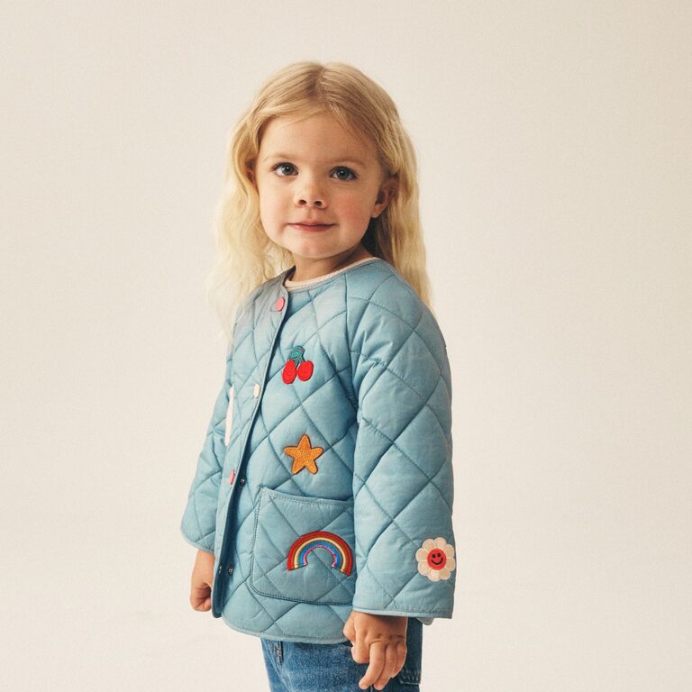 A young girl with long blonde hair wears a pale teal-green quilted jacket with embroidered cherry, star, rainbow, and flower badges, over blue jeans against a cream backdrop.