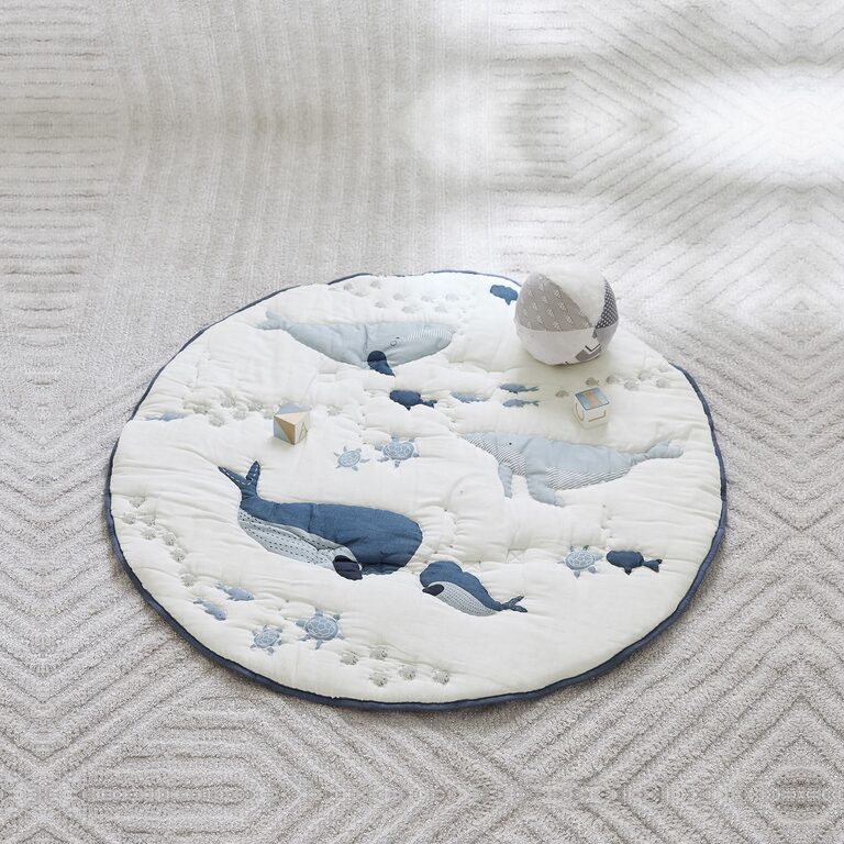 A round, quilted ivory muslin play mat appliqu&eacute;d with blue whales and small sea turtles and edged in navy, laid on a pale grey textured rug with a fabric ball and two wooden blocks.