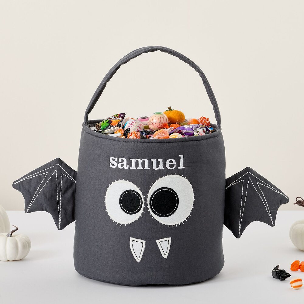A charcoal-grey bat treat bag filled with Halloween candy, with round white felt eyes, two white felt fangs, stitched bat wings at the sides and the name &ldquo;samuel&rdquo; embroidered above the face.