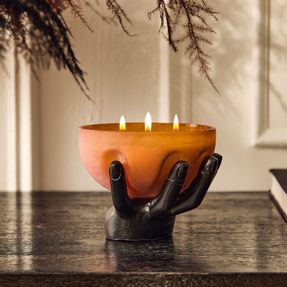 A lit three-wick candle in an amber glass bowl cradled by a black sculpted hand, on a dark wood mantel beneath trailing dried branches.